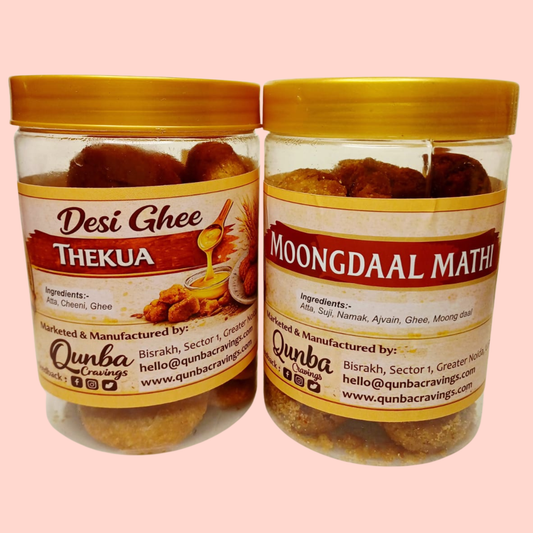 Two transparent snack jars from Qunba Cravings placed side by side, one containing traditional Thekua and the other Moong Daal Matthi, both with red lids and ornate cream-and-gold stickers featuring the Qunba Cravings logo, family illustration, product names, and homemade Indian snack branding.